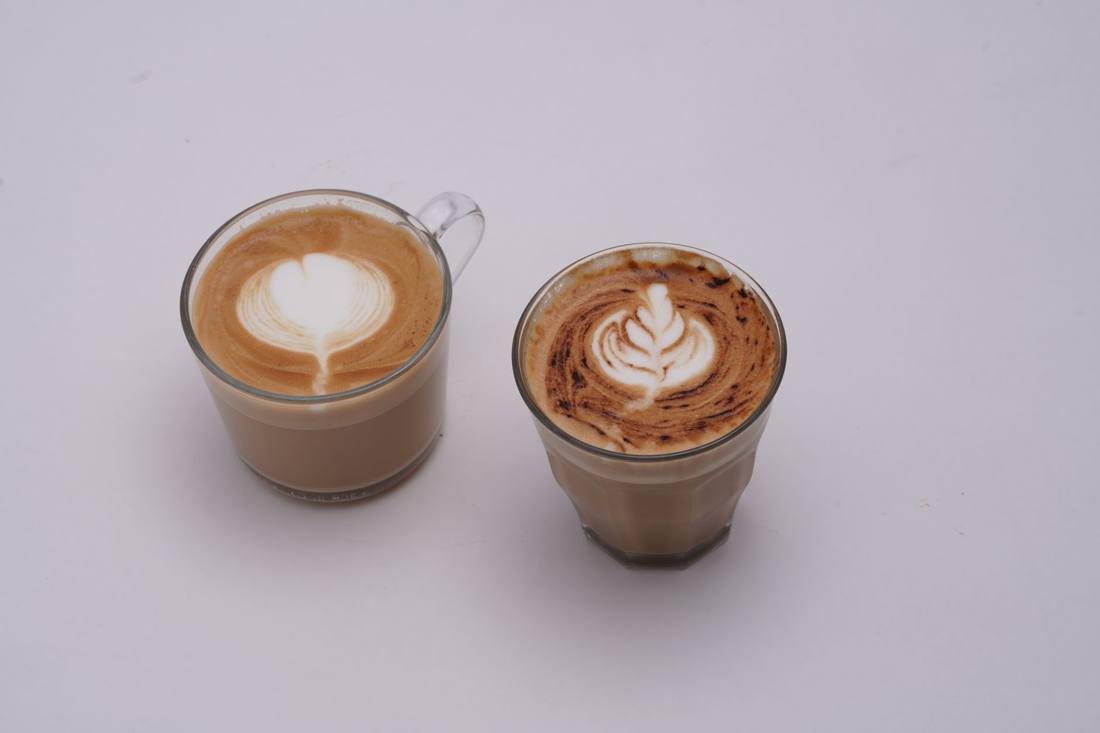 Two glass cups of latte with intricate latte art on white background.