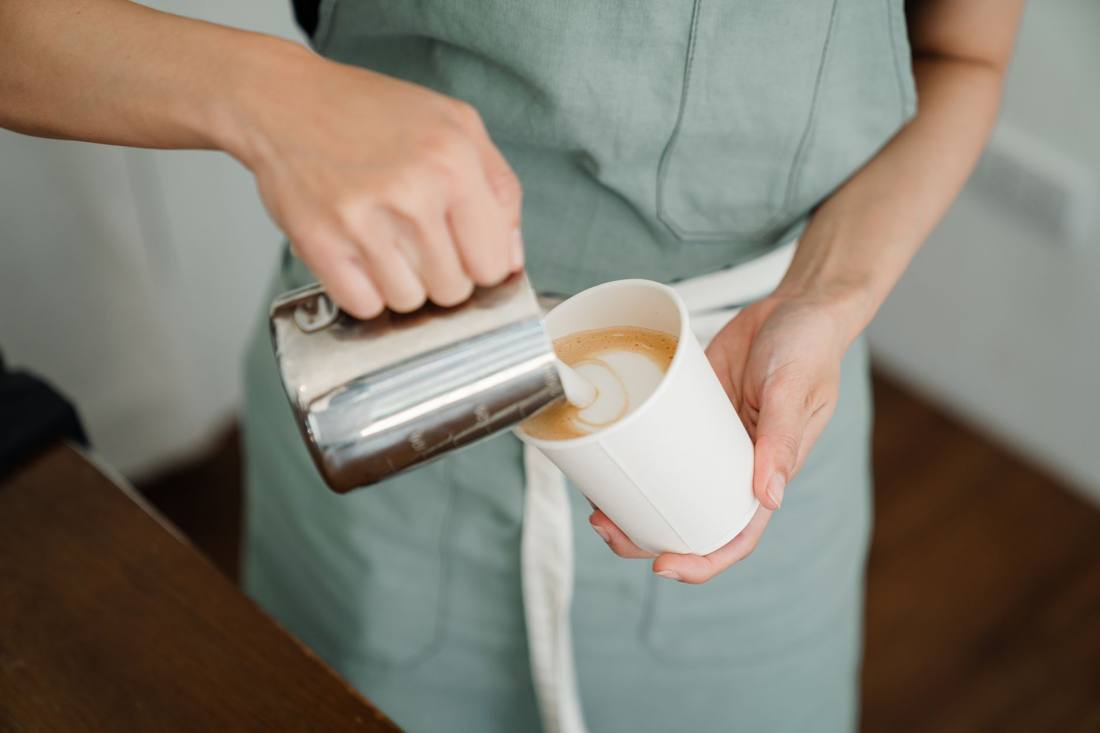 Unrecognizable female in apron pouring hot milk into paper cup while preparing aromatic latte to go in modern coffee house